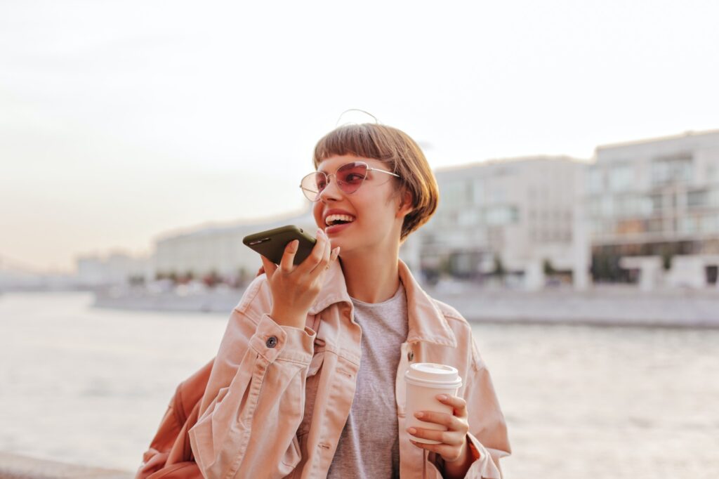 Stylish woman posing with phone in city. Short-haired teen girl in pink glasses and beige jacket sm
