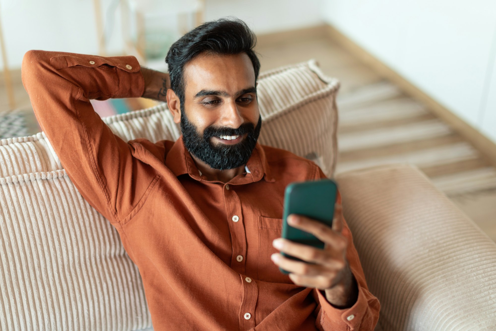 Cheerful Indian Bearded Guy Texting Using Phone In Living Room