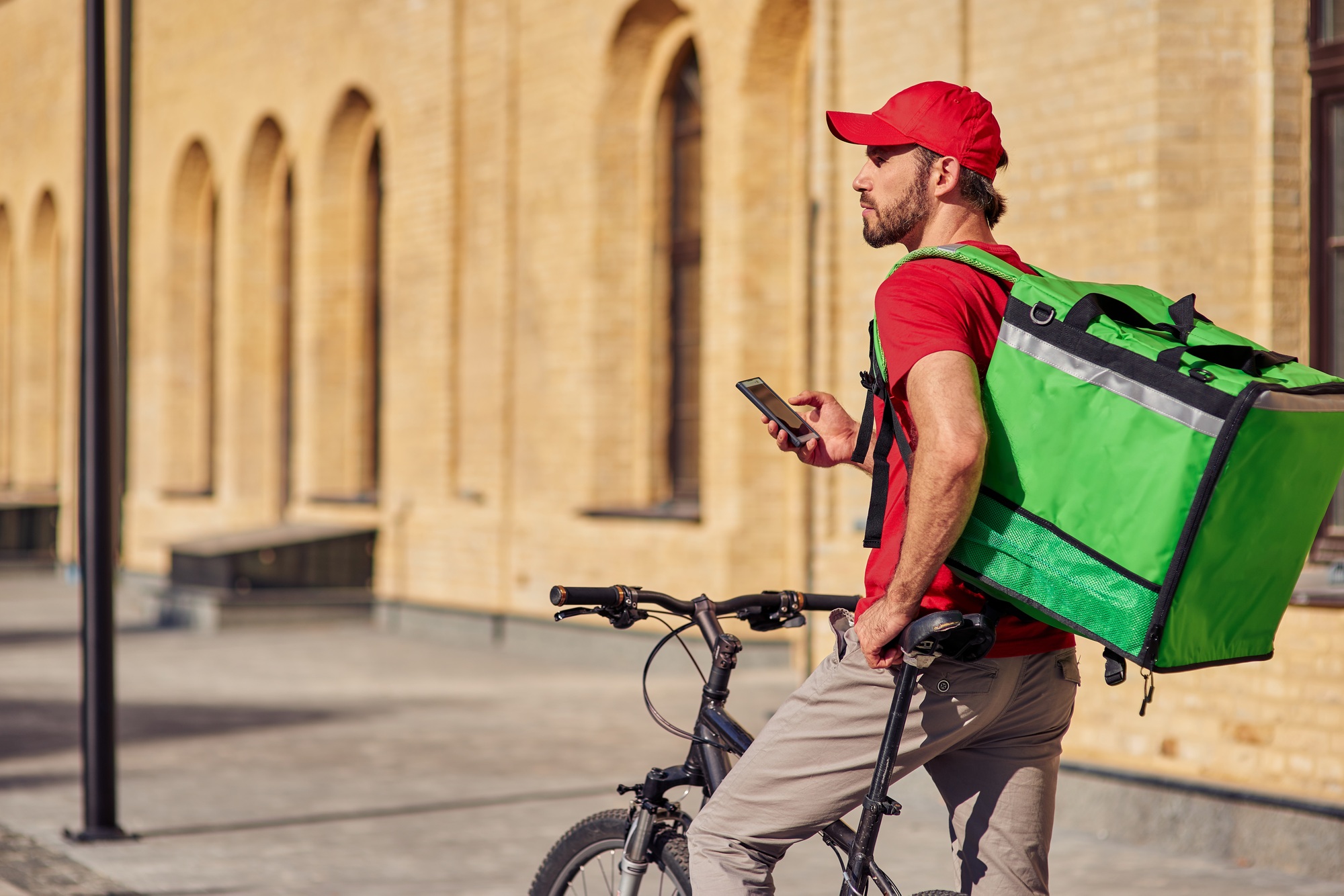 Tracking order. Young male courier with thermal backpack using mobile app while standing on the