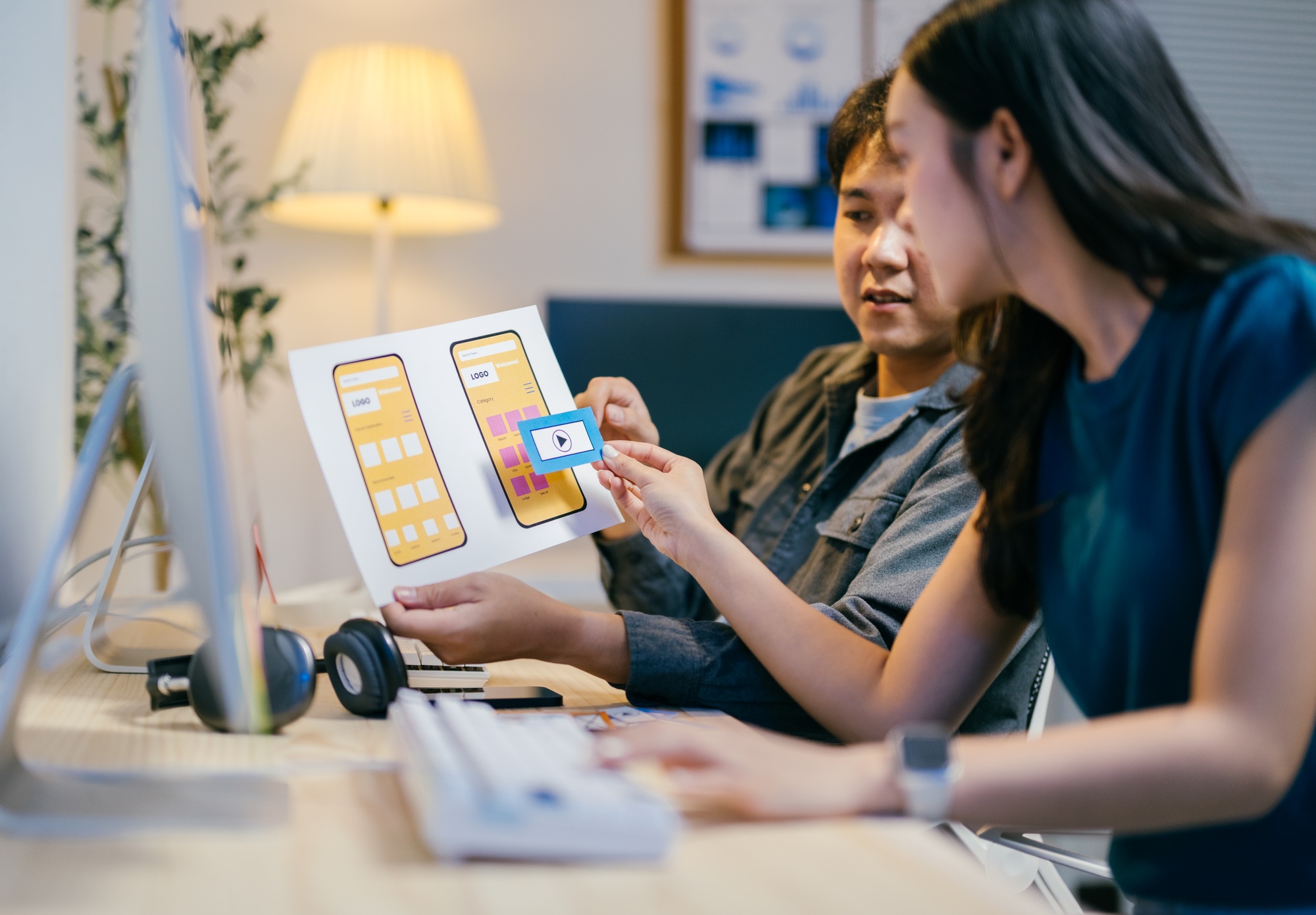 Two asian ui/ux designers are discussing a mobile app wireframe prototype at their desks in the offi